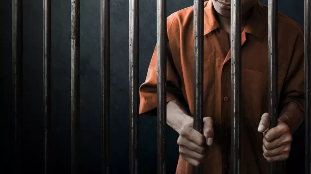 Narapidana mengenakan pakaian tahanan berdiri di balik jeruji penjara.