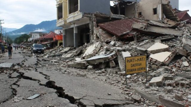 Bangunan rumah rusak berat dan jalan retak akibat gempa, dengan puing-puing bangunan berserakan di kawasan terdampak.