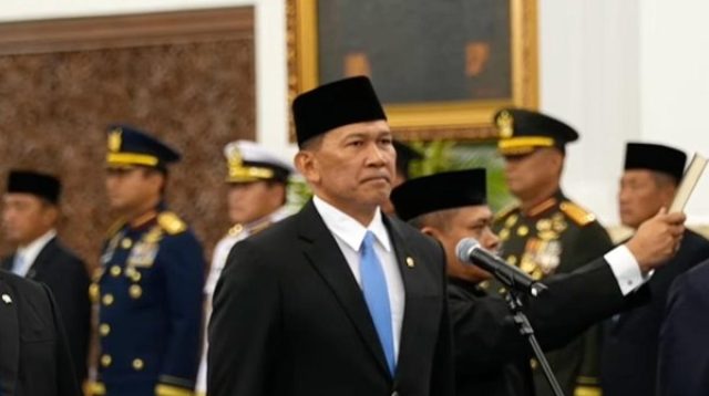 Seorang pejabat negara mengenakan jas hitam, dasi biru, dan peci hitam saat mengikuti prosesi pelantikan di Istana Kepresidenan, dengan sejumlah pejabat berseragam militer di belakangnya